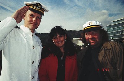 Three individuals pose outdoors in formal naval and casual attire, likely mid-1990s. The man on the left wears a white naval ...