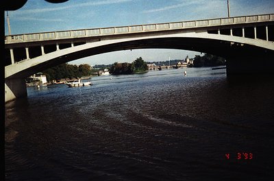 Vintage archival shot of a concrete bridge spanning a river, framed by its own arch. Mid-20th century urban landscape with bo...
