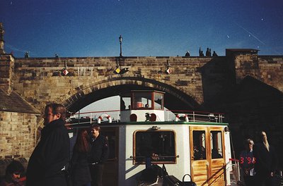 Historic boat tour beneath Prague Castle’s stone archway, 1990s. Wooden and glass-covered vessel with passengers enjoying vie...