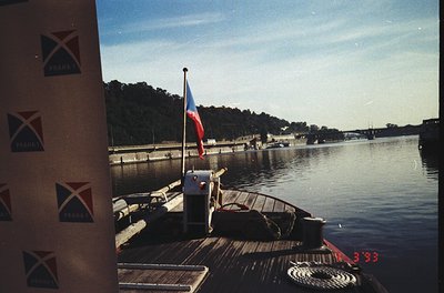 Vintage boat docked at Prague’s Vltava River, 1993. Czech flag flying, "PRAHA" branding on wooden pier. Urban greenery and hi...