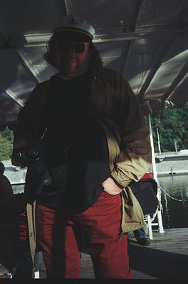 Vintage photo of a man in a naval officer’s peaked cap, sunglasses, and red trousers aboard a ship. Dark coat, beige jacket, ...