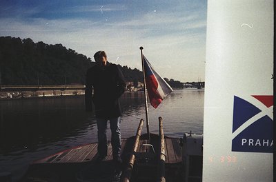 Vintage boat docked at waterfront with individual in dark jacket and bell-bottoms, holding a flag featuring red, white, and b...