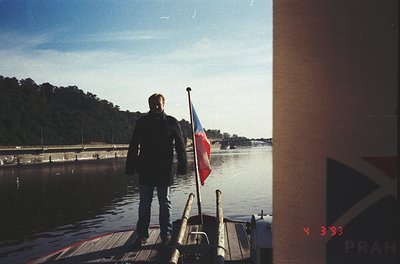 Vintage photo of a man standing on a wooden dock, holding a flagpole with the Czech Republic flag. Industrial waterfront with...