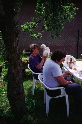 Two women relax outdoors in a shaded garden setting, likely mid-1990s. One holds a small white dog, while the other sits with...