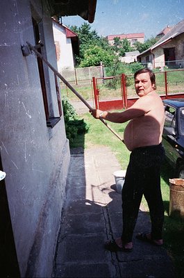 Man in mid-swing with a long wooden pole, likely a traditional *koprižnik* (pole-dancing tool), in a residential courtyard. W...