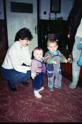 A woman in a white blouse and blue jeans kneels beside two young children in a dimly lit indoor setting, likely a 1980s-1990s...