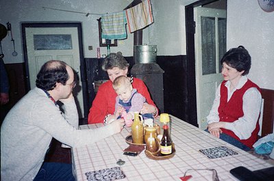 Family gathering in a modest kitchen, 1970s-1980s Eastern Europe. Three adults and a toddler seated around a table covered wi...
