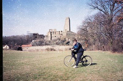 Vintage photo of a person in cycling attire riding a basic frame bike near a medieval stone ruin with a tower and arched wind...