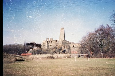 Vintage photo of a ruined medieval tower and stone walls, likely Eastern European. Overgrown grass and a soccer goal in foreg...