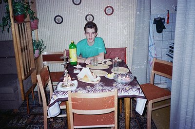 Vintage indoor dining scene featuring a young person seated at a wooden table covered with a floral-patterned tablecloth. Sur...