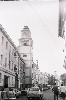 Black-and-white street scene featuring a prominent **clock tower** with a domed roof, flanked by **1960s-era buildings** in a...