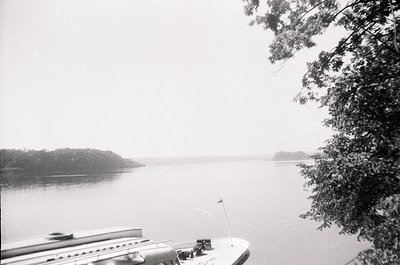 Mid-20th century black-and-white lakeside scene with misty horizon. A docked boat’s edge and flagpole frame the shot, while a...