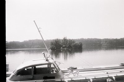 Mid-20th century boat docked at a serene waterfront, likely a lake or coastal inlet. The vessel features a tall mast with a f...