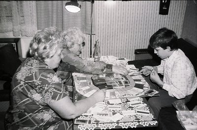 A mid-20th century indoor scene showing two adults and a child engaged in crafting. Spread across a table are paper cutouts, ...