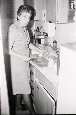 Mid-century kitchen scene featuring a woman in a patterned apron preparing food on a vintage countertop with a manual coffee ...