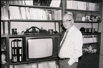 Mid-20th century man in a bookstore/library setting, posing beside a vintage CRT TV displaying static. Surrounding shelves ho...