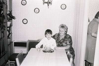 A black-and-white indoor family moment: a young boy and an older woman pose at a table with a lit birthday cake. The woman we...