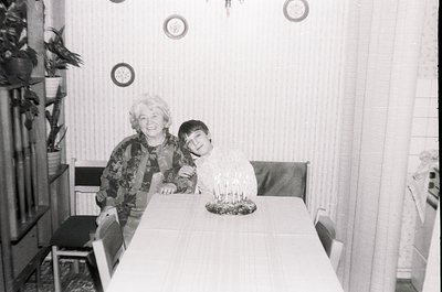 A mid-20th century indoor portrait showing a woman and young boy seated at a simple wooden table. The woman wears patterned c...