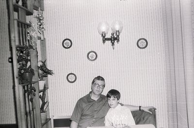 Mid-century indoor portrait: man and boy seated on a striped sofa in a modestly decorated living room. Wallpaper features cir...