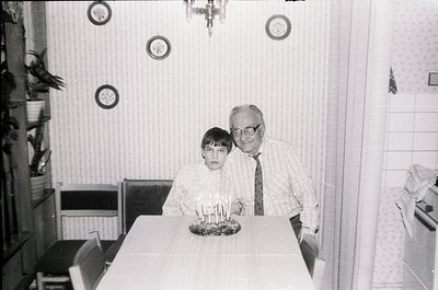 Mid-20th century indoor family portrait: man in suit and tie poses with young boy beside a birthday cake with lit candles. Pl...