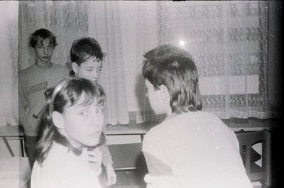 Vintage black-and-white photo of four children in a dimly lit indoor setting, likely a classroom or home. One child wears a "...