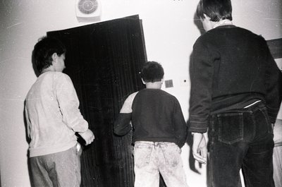 Three individuals in a dimly lit indoor setting, likely a 1970s–1980s home or office. The central figure wears a long-sleeve ...