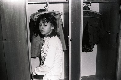 Black-and-white shot of a young individual in a narrow closet, holding a hat above their head. The person wears a long-sleeve...