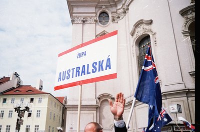 Baroque-style church façade with ornate columns and arched doorway. Crowd holding a red-and-white banner reading "Župa AUSTRA...