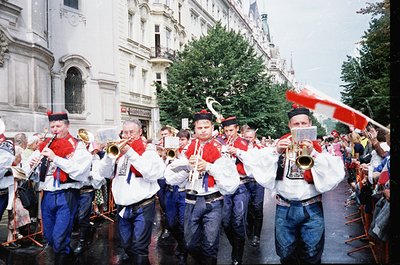 Uniformed brass band performing in a European city street, likely 1980s–1990s. Members wear white jackets with red accents, b...