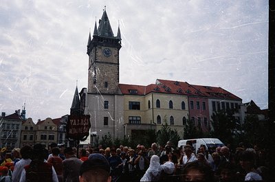 Historic clock tower with Gothic Revival architecture dominates a crowded public square. Crowd gathered beneath a banner read...