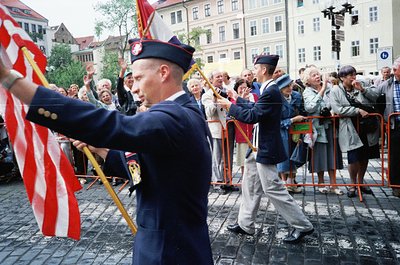 Uniformed marchers in formal attire carry flags during a public parade, likely in a European city square. The attire suggests...