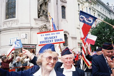 Crowd of elderly men and women in blue uniforms and red caps march in a parade, holding flags and a banner reading "D.A. Soko...