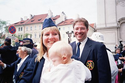 Historic parade scene in Prague’s Old Town, featuring a woman in a blue cap holding a baby, flanked by a man in formal attire...