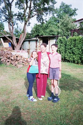 Family portrait in a rural backyard, 1990s. Three individuals pose with a tennis racket, surrounded by a stone wall and woode...