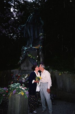 Couple sharing a tender moment at a solemn monument adorned with floral tributes. Dark, dramatic lighting enhances the statue...