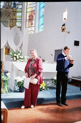 A woman in a maroon dress and patterned blouse holds a small booklet, standing beside a modernist altar adorned with white fl...