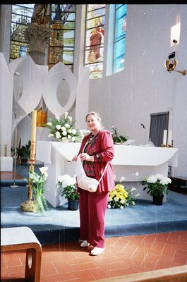 Woman in maroon suit holding a small white clutch stands at a modernist altar in a church, surrounded by floral arrangements ...