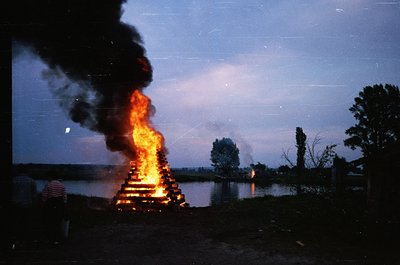 Vibrant bonfire at dusk beside a reflective body of water, surrounded by silhouetted trees and low-lying structures. Smoke ri...