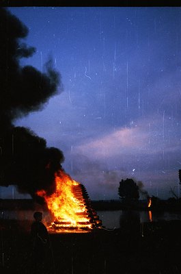 Vibrant nighttime bonfire under rain, silhouetted figures observing. Flickering flames illuminate a wooden structure, likely ...