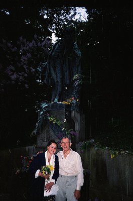 Couple posing beside a dark, moss-covered stone monument with floral tributes, likely a war memorial. Man in light-colored sh...