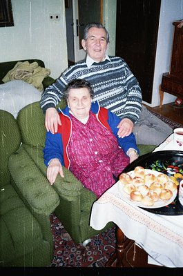 Couple posing indoors on a floral-patterned sofa, likely late 20th century. Man in striped sweater, woman in red-and-blue jac...