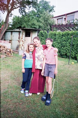 Family portrait in a residential backyard, likely late 20th century. Three individuals pose outdoors near a weathered shed an...