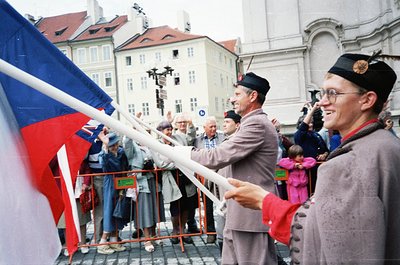 Two men in traditional Czech folk attire—wide-brimmed hats and embroidered vests—hold a large Czech flag during a public gath...