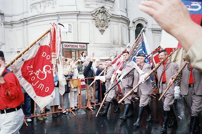 Traditional folk dancers in period costumes perform in a European city square, holding flags with Cyrillic script ("ДОНЖО") a...