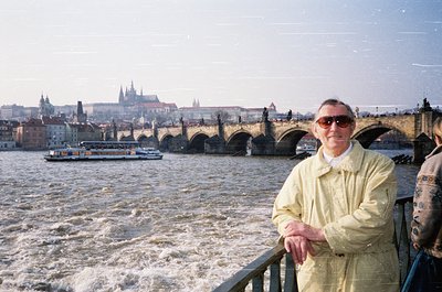 Man in light beige jacket poses by Prague’s Charles Bridge, with the Vltava River’s rapid flow below. Gothic spires of Prague...