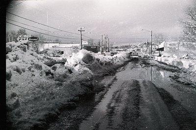 Mid-20th century street blanketed in heavy snow, likely post-1950s. Snowbanks line both sides of a partially cleared road, wi...