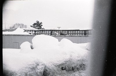 Mid-century industrial scene framed by heavy snowfall, likely 1950s–1960s. Snowdrifts obscure a low-lying building with a cor...