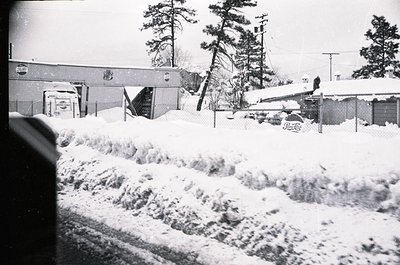 Black-and-white winter scene showing a snow-covered residential area with a partially buried road. Snowdrifts line a street, ...