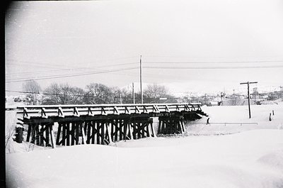 Black-and-white winter scene of a wooden truss bridge spanning a snow-covered stream. Snow blankets ground, trees, and bridge...