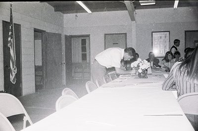 Mid-century indoor gathering with floral centerpiece on a long table. A man in a white shirt bends to place a wreath on a pla...